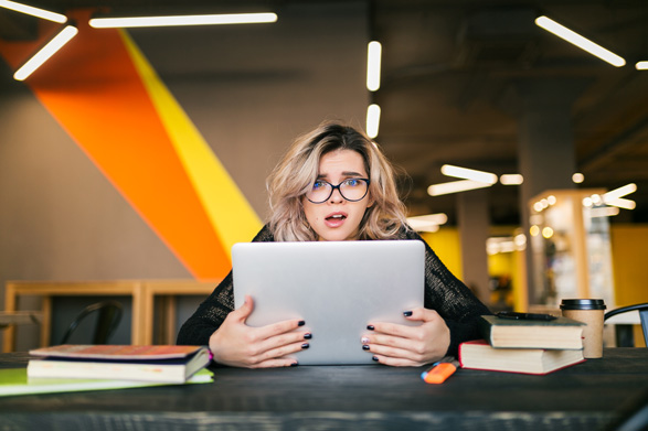 portraet-der-jungen-huebschen-frau-mit-gesockeltem-gesichtsausdruck-sitzend-am-tisch-der-am-laptop-arbeitet